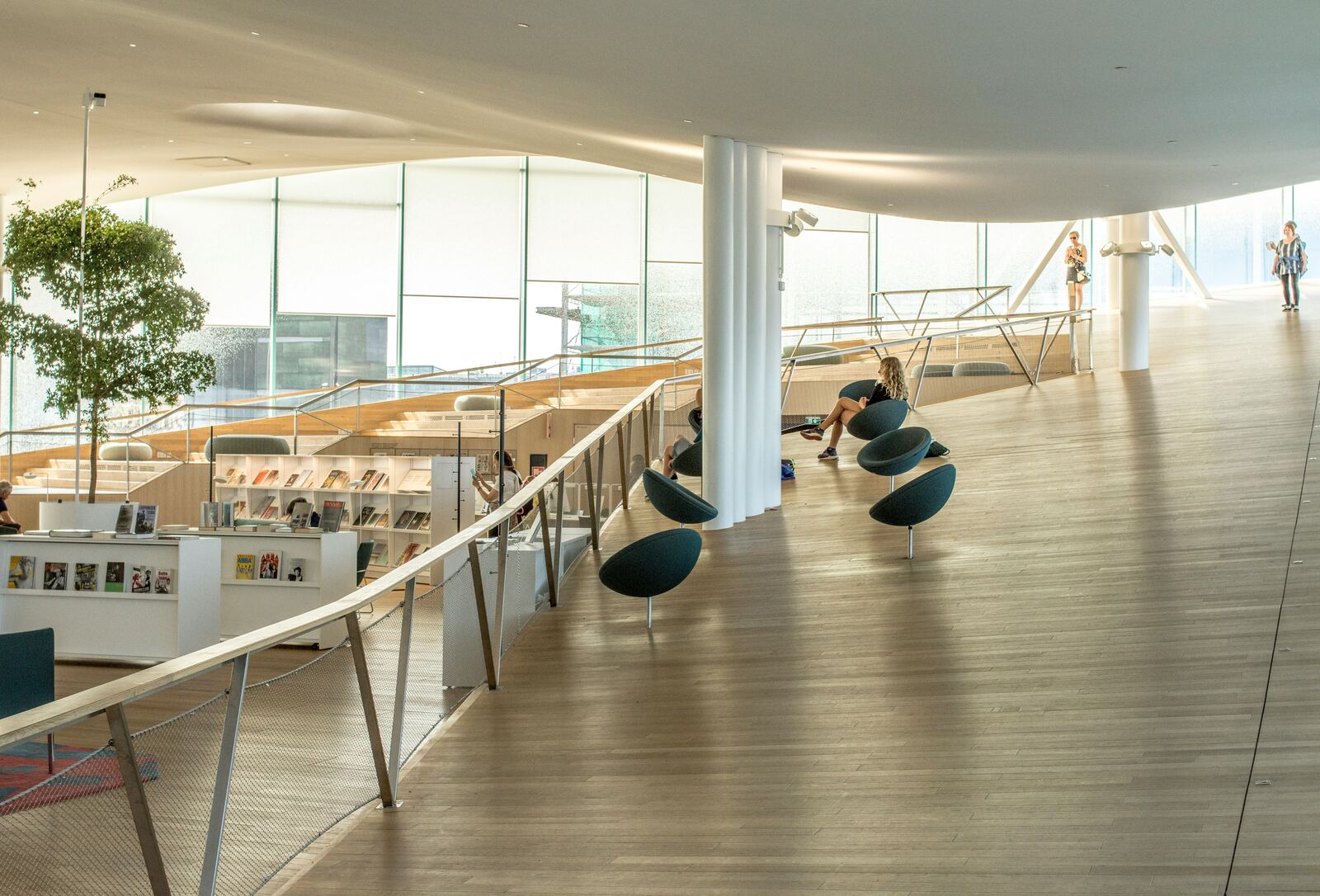 Adults reading in the warm afternoon light of a bright Nordic library hall, beneath a sweeping white ceiling.